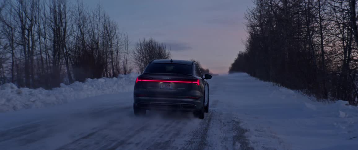 A black car with tail lights on travels on a snow covered road lined with trees