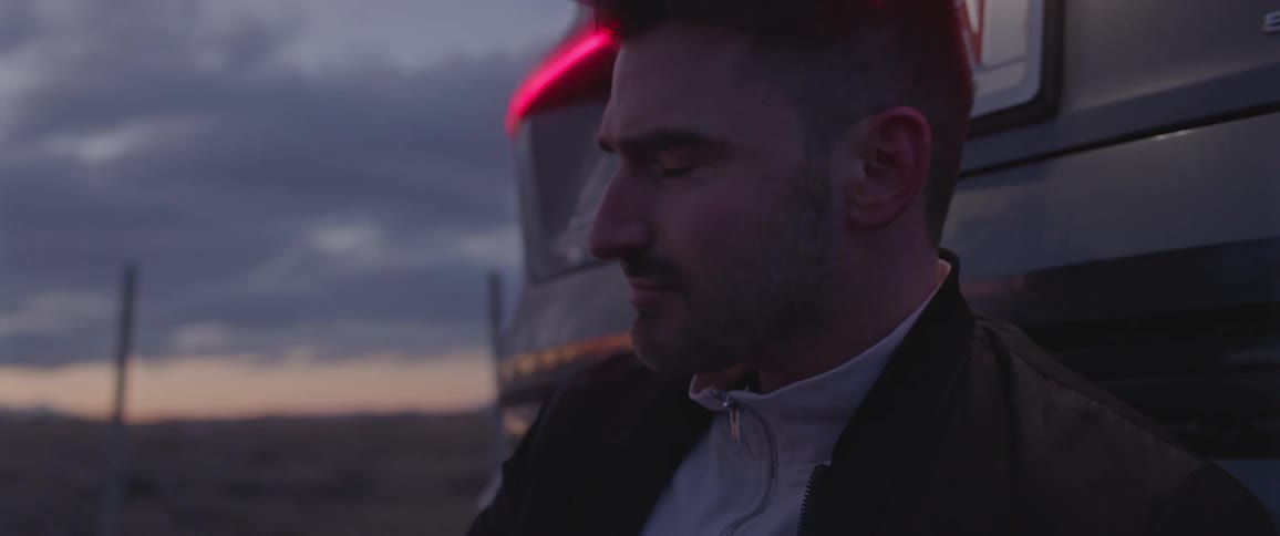 A man sitting on the ground leans against the back of his car and looks around