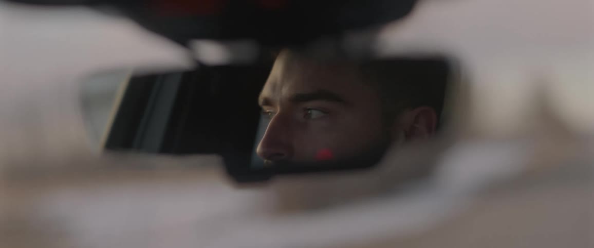A reflection of a man driving a vehicle in the rearview mirror with trees the sky and clouds in the background
