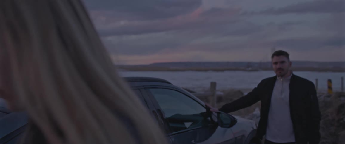 Two people stand outside parked car looking at each other and away on snow dotted shore with red tinted sky in background
