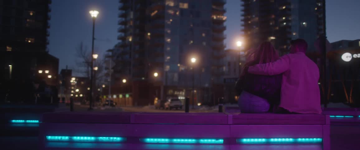 A man and woman sit closely on a lit bench outside in a large city