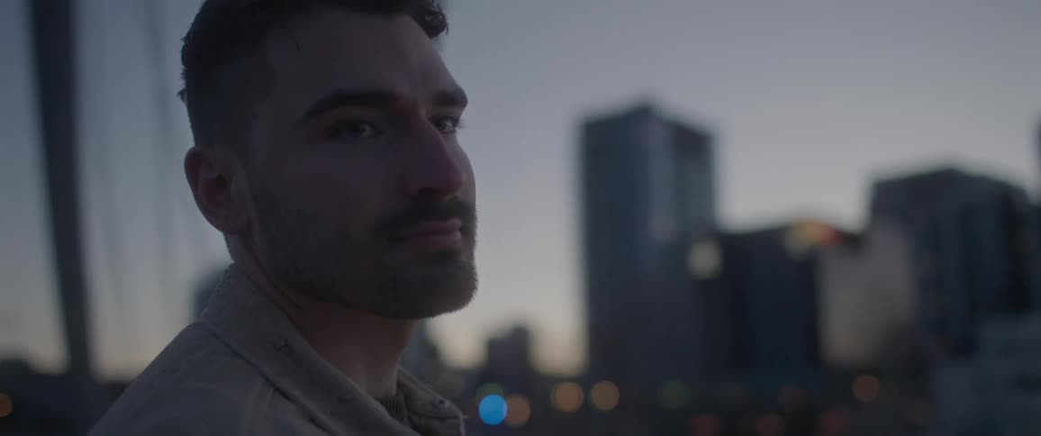 Man with a brown jacket and a stubble beard looks at his girlfriend on the bridge at dusk