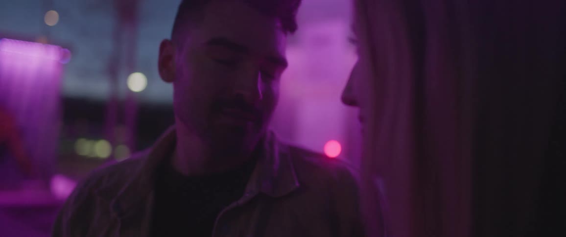 A man leans in to kiss a woman who stops him with a hand as they sit together outdoors at night