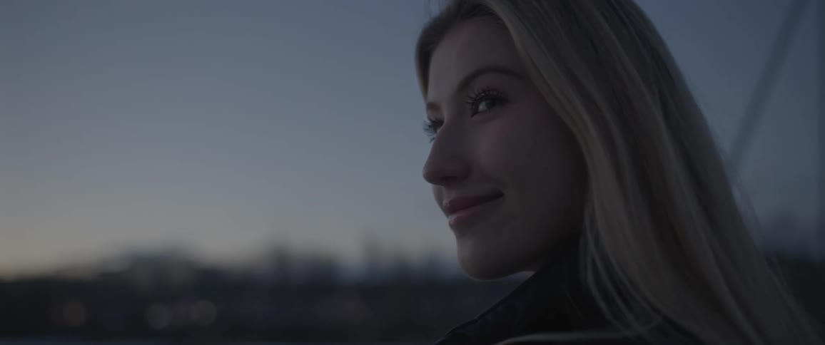 Blonde woman stands outside across from leafless trees smiling on windy day with red tinted light horizon in background