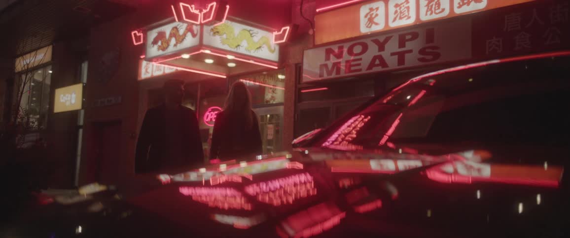 Couple walks out of restaurant to car reflecting neon signs