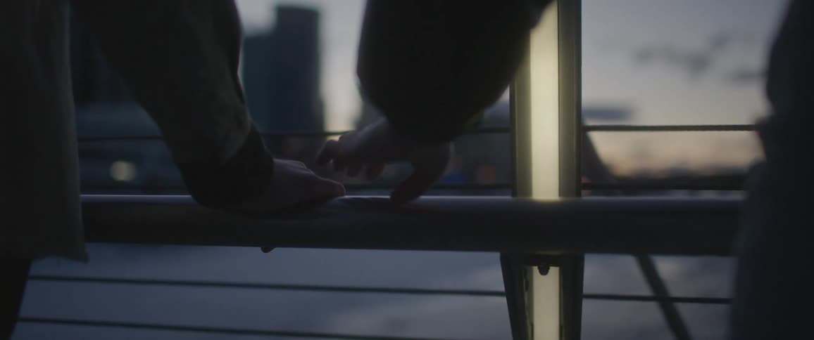 Two people standing on a bridge near the city hold hands at sunset