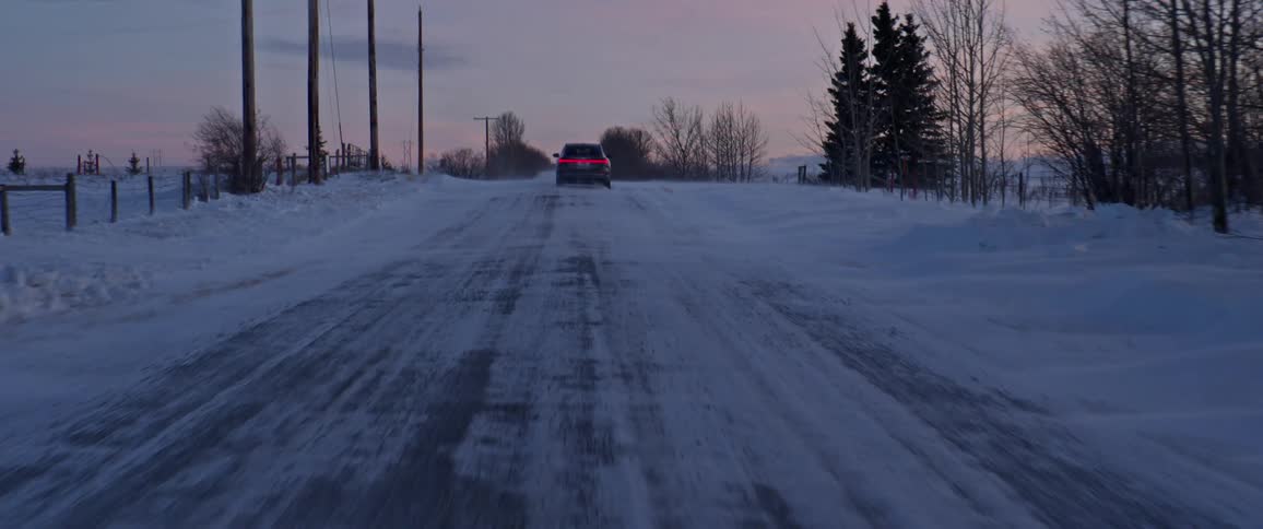 A black car travels on a snow covered road with trees along the roadside
