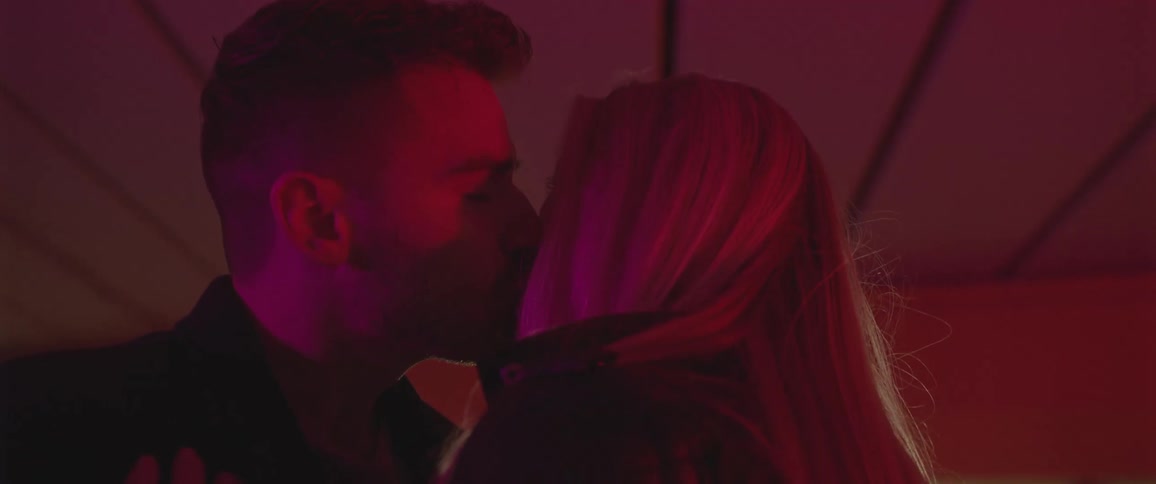 Man with a beard and blonde woman with a black leather jacket kiss each other on lips in red light