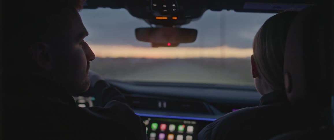 A driver and passenger glance silently at one another as they travel down a rural road in a vehicle in the evening