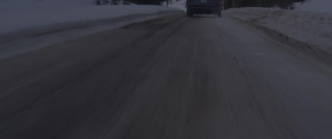 A vehicle driving on a snowy road with trees a mountain range the sky and clouds in the background