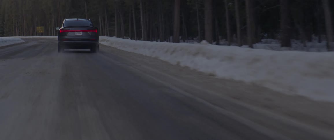 A dark colored sedan is driving down a wet road surrounded by snow and a forest