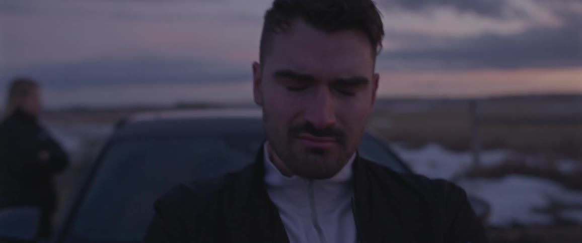 A man shakes his head in disappointment while sitting against the hood of a car parked on the side of a rural road at sunset