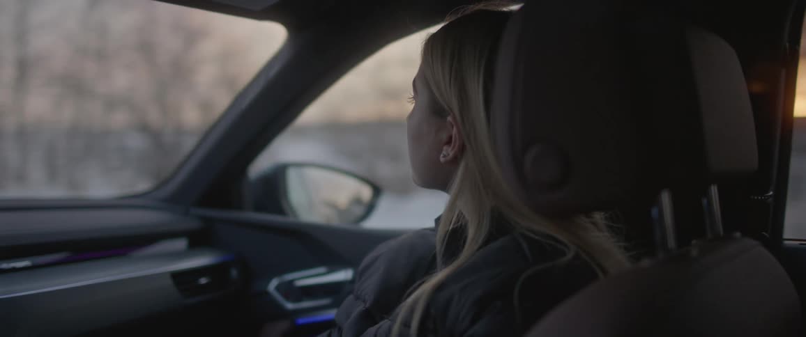 Blonde woman sits in passenger seat as car drives past leafless trees and snow covered fields while sunlight glows on the horizon