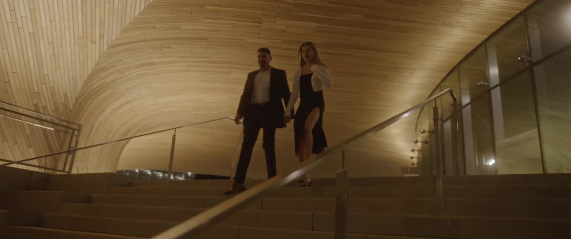A man and woman walk out of a large building at night and hold hands walking down a staircase