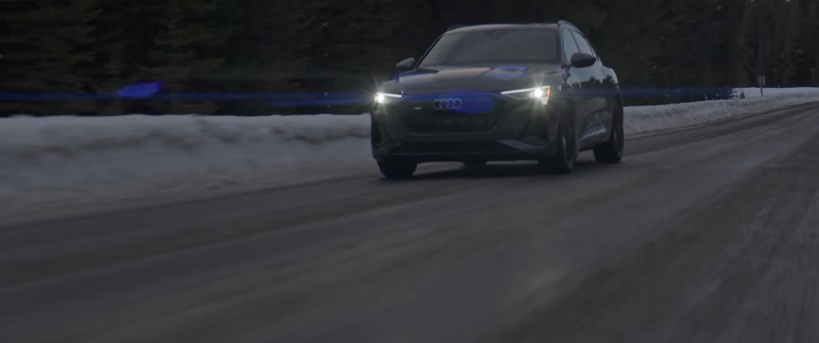 A black car with headlights on travels on a road with snow along the roadside