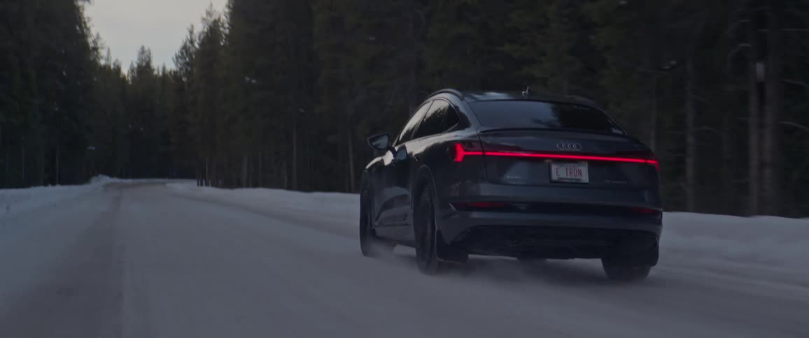 A black car travels on a snow covered forest road lined with tall trees