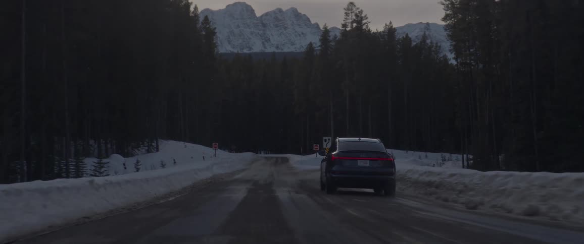 A vehicle turns onto an exit ramp while driving on a snow covered road surrounded by trees and near a mountain range