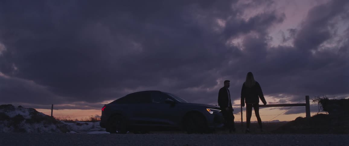 Two people talk near car parked near mounds covered in snow under a cloudy sky