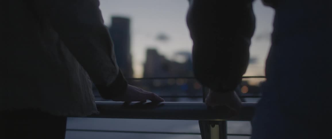 Couple slide hands across rail to touch standing across water from city skyline backed by glowing horizon