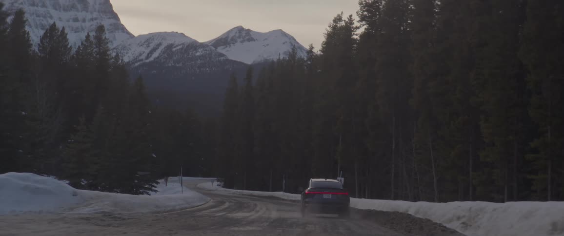 A black car travels on a snow covered road with trees along the roadside and snow covered mountains in the distance