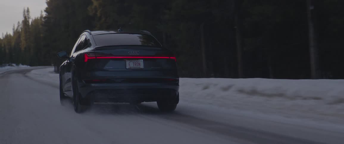 An audi vehicle travels on a snow dusted rural road surrounded by tall trees