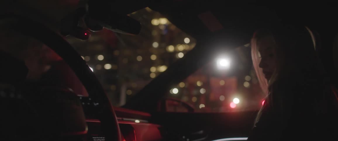A woman sitting in a vehicle with a steering wheel with a man and buildings in the background