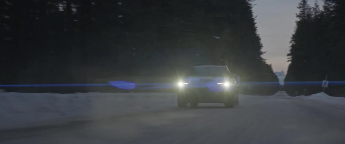A black car travels on a snow covered road with headlights on and trees along the roadside