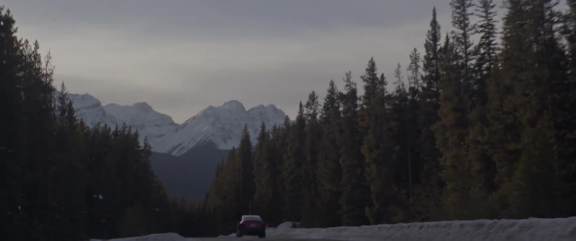 Black car driving on a snow covered road throught the forest near the mountains