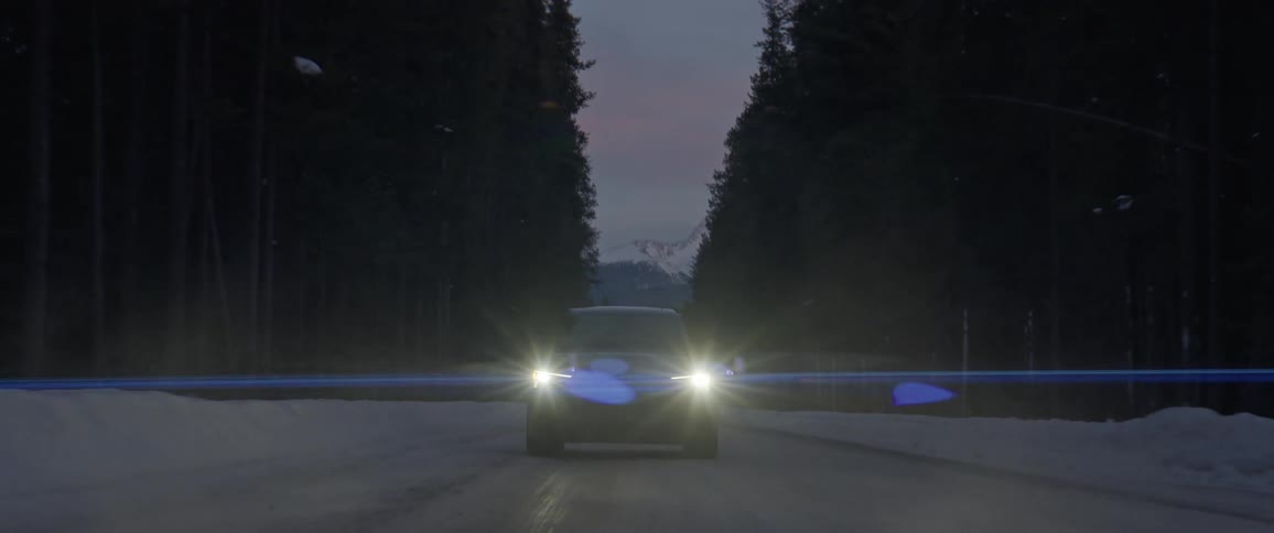 A car with headlights on travels on an ice and snow covered road surrounded by tall trees