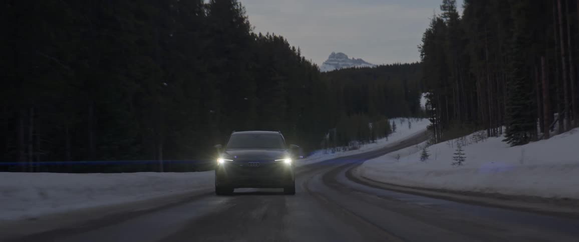 A black car with headlights on travels on a road lined with trees and snow