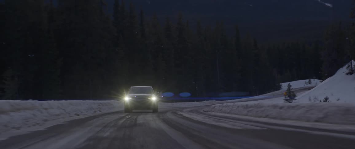 A black car is driving on a road with headlights on during snowy season