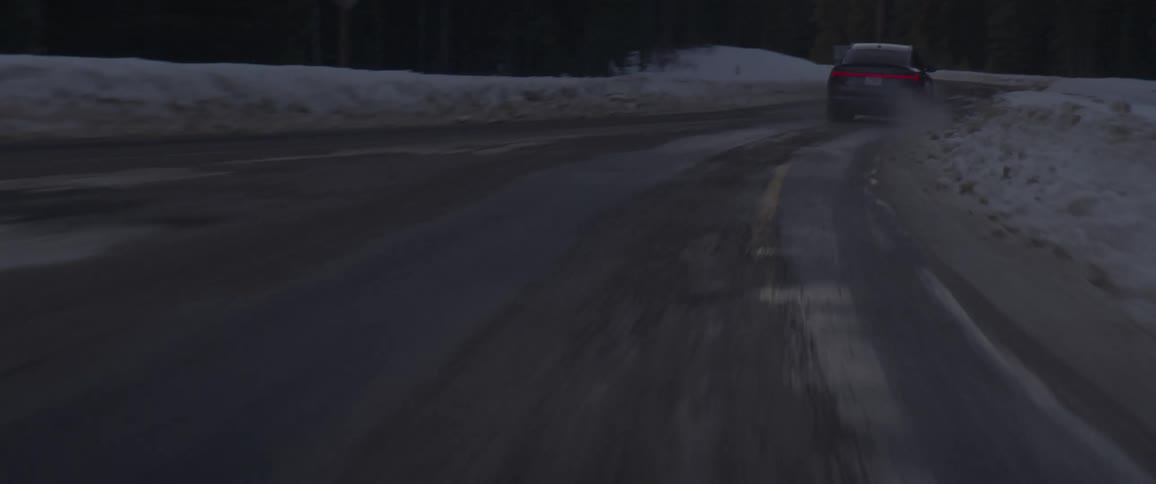 A black car travels on a two lane road with snow along the roadside
