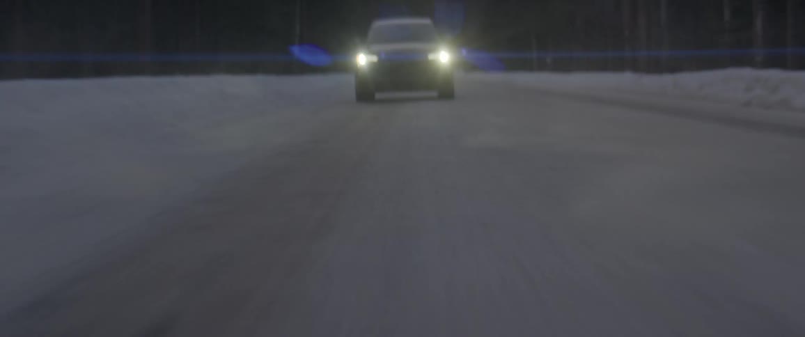 A black car with headlights on travels on a snow covered road with trees along the roadside