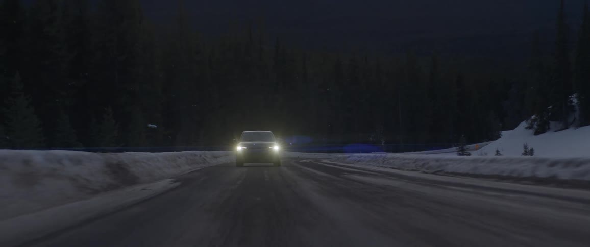 A vehicle with headlights driving on a snowy road with trees and a mountain range in the background