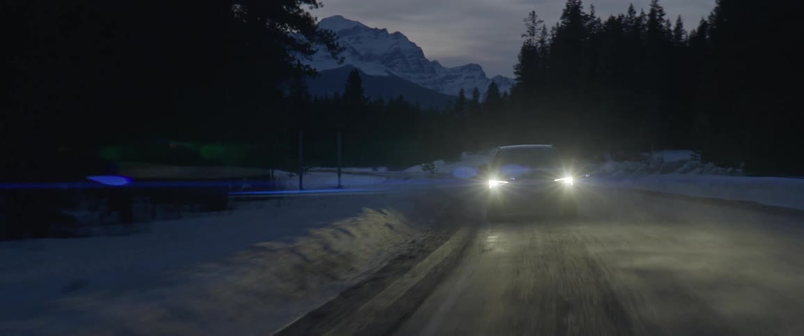 A vehicle travels along a wintry road through a forest in the evening