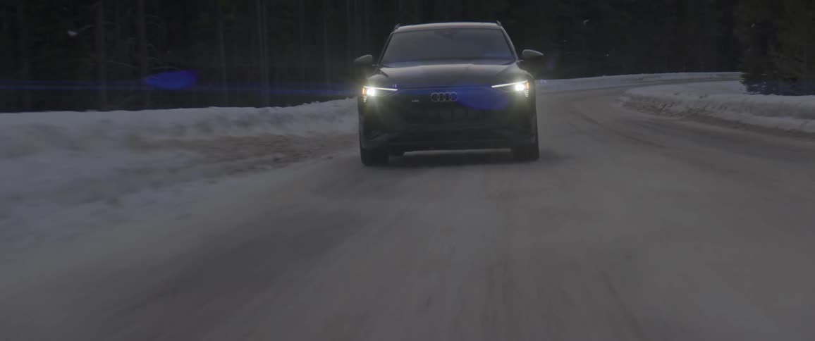 A black automobile with headlights on travels on an ice and snow covered road lined with trees