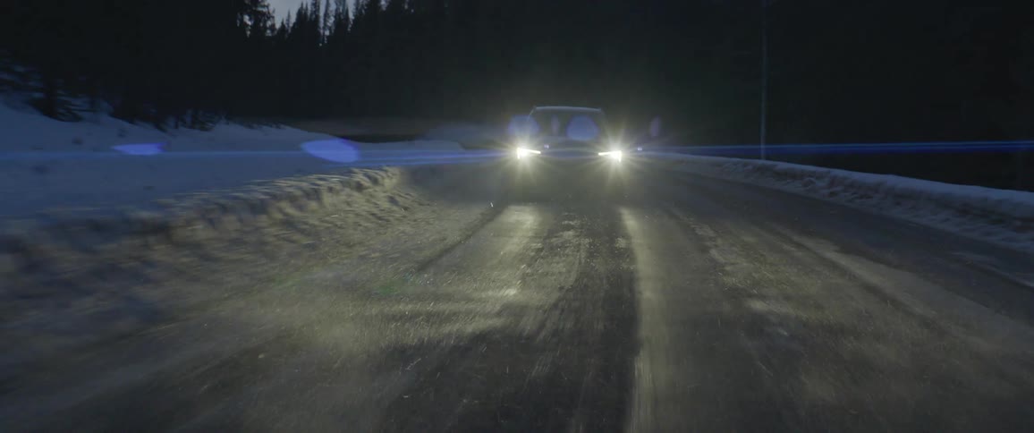 A vehicle travels along a road through an evergreen forest on a winter evening