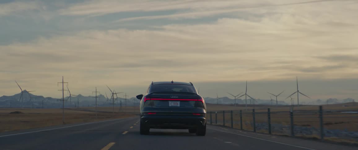 Dark grey audi car drives on highway with wind turbines in the background
