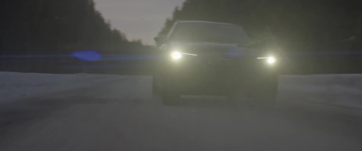 A car with its headlights on speeds along a snowcovered road that is lined with forests