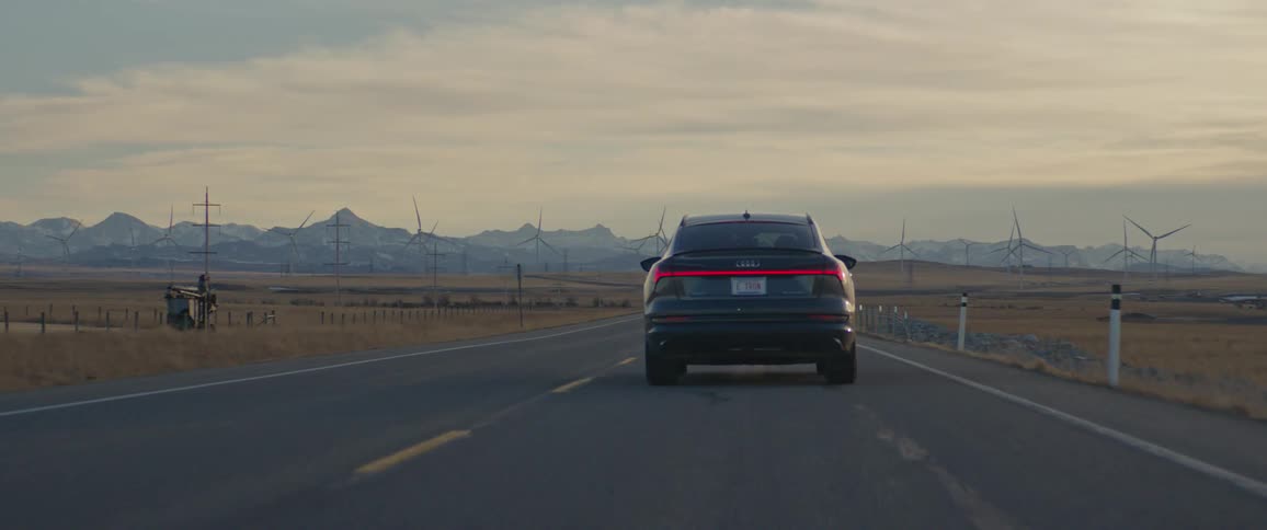 A black car travels on a road with utility poles along the roadside and wind turbines in the distance