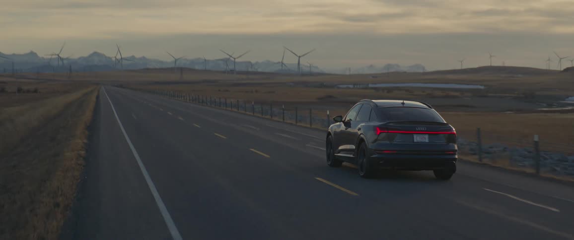 A black car travels on a two lane road with spinning wind turbines in the distance