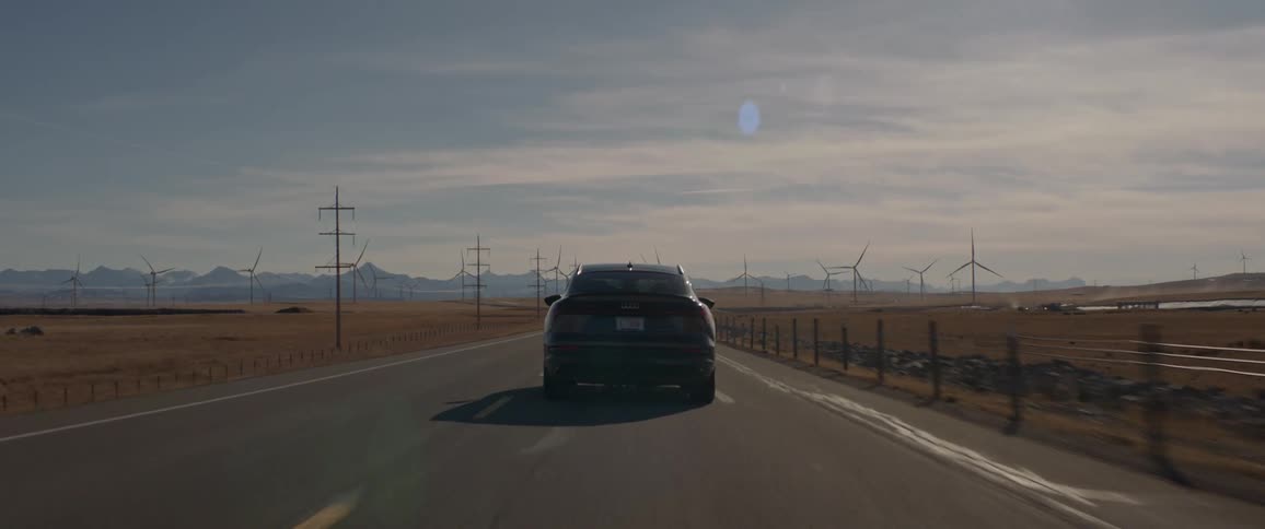 A black car travels on a road with utility poles along the roadside and spinning wind turbines in the distance