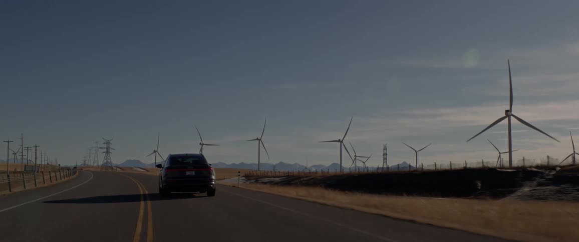 Dark grey audi car drives on highway next to wind farm