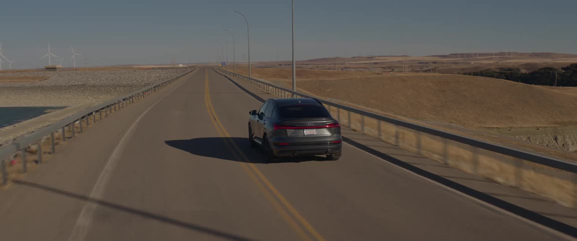 A car travels on a road with utility poles along the roadside and spinning wind turbines in the distance