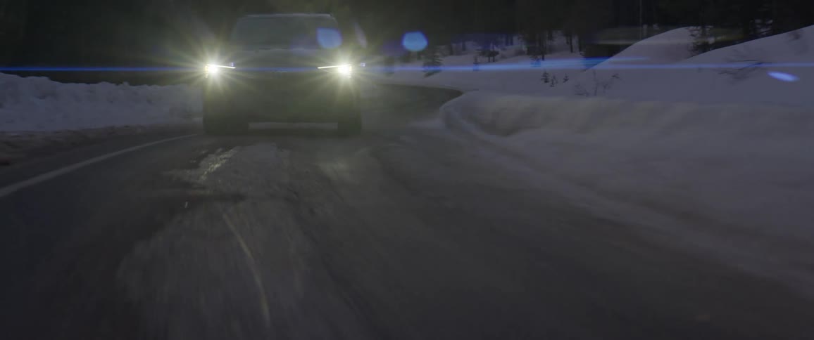 A vehicle with headlights driving on a snowy roads with trees in the background