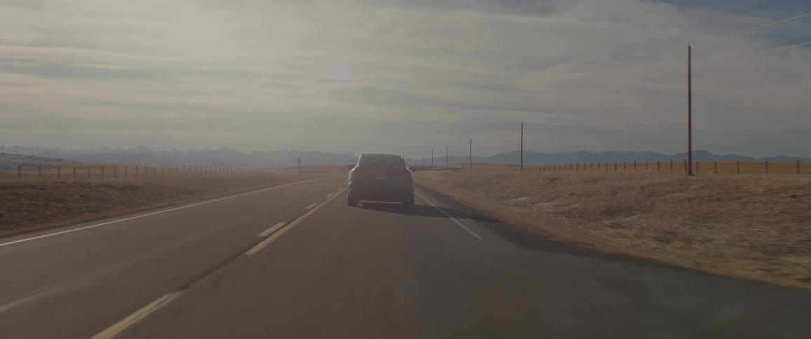 A black car travels on a road with utility poles along the roadside under a cloudy sky
