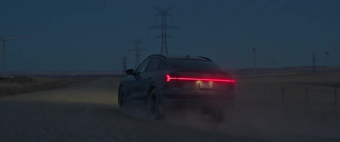 An automobile drives along a dusty road past wide open fields