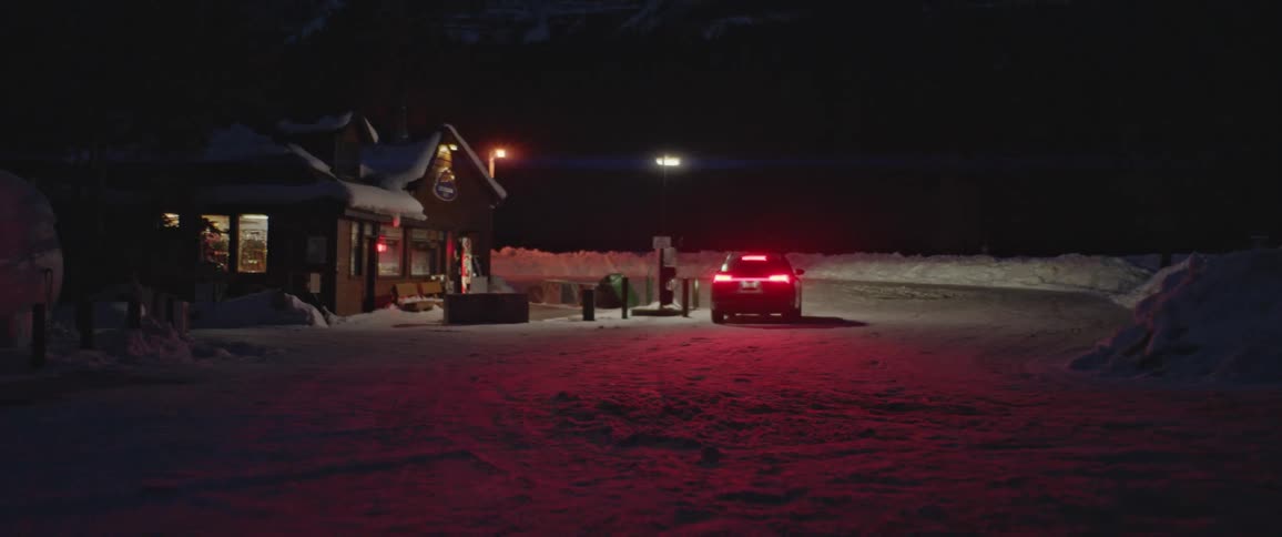 A vehicle driving on a snowy road next to a store with the sky and clouds in the background