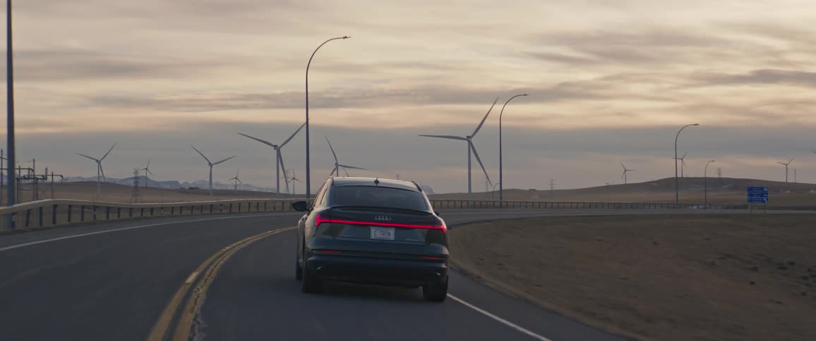 Dark gray audi car drives on highway next to wind farm at dusk