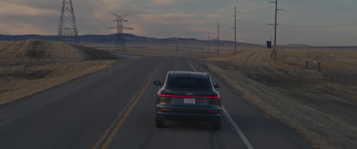 A black car travels on a two lane road with utility poles and utility towers along the roadside